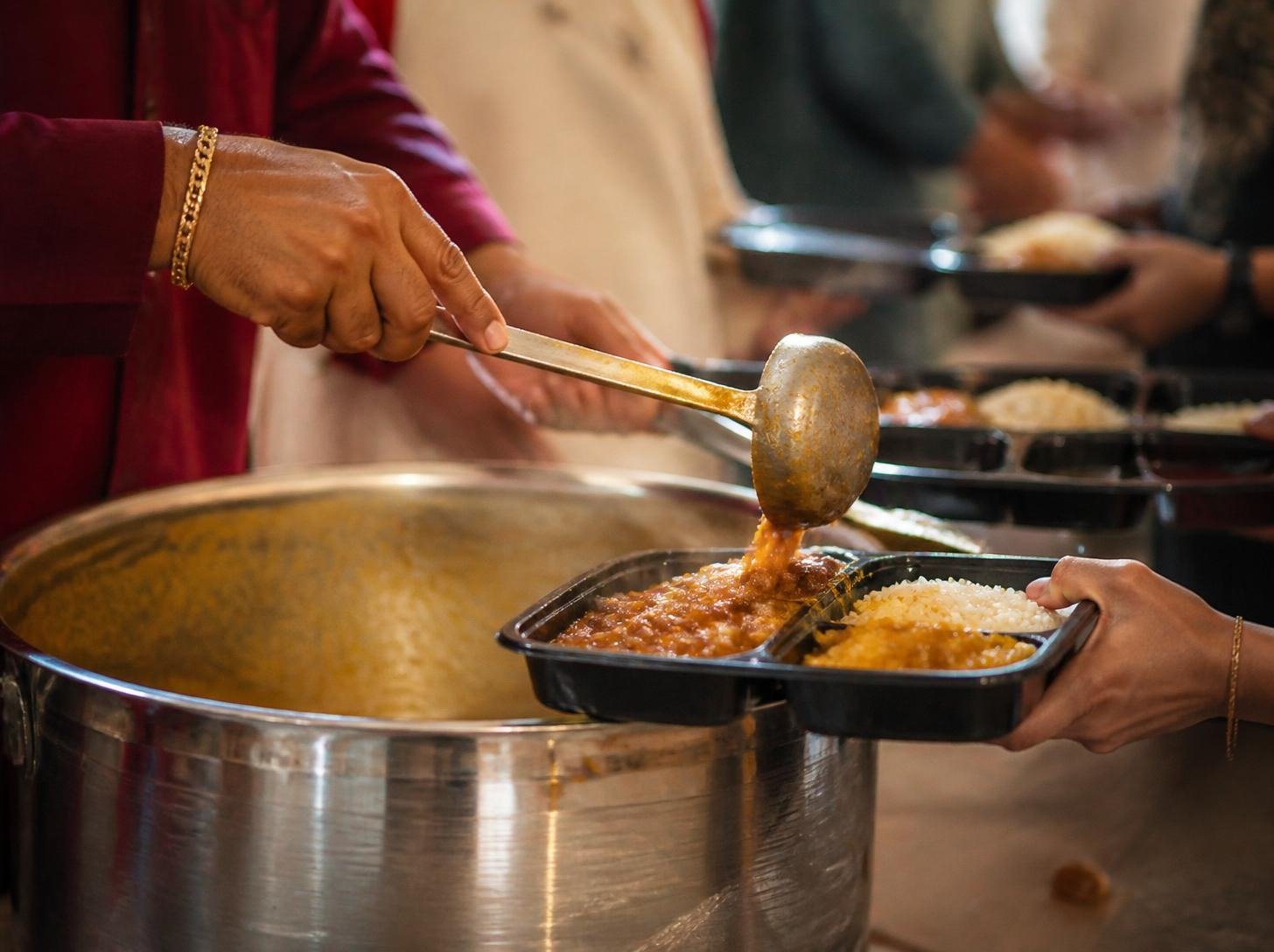 Volunteer hands serving food from a large pot into a langar tray